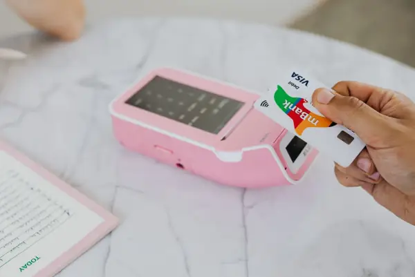 Hand tapping a Visa card on a pink contactless payment terminal